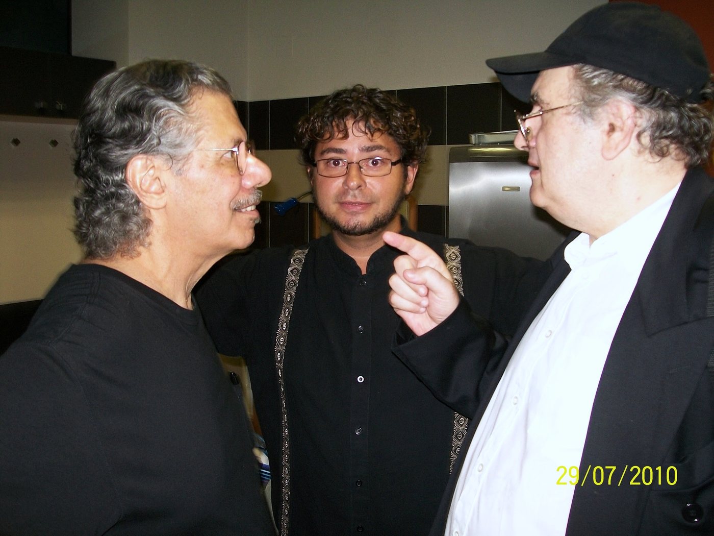 Yan Kayary entre dos hombres, uno de cabello gris ondulado y otro de gorra negra que le habla señalando con la mano