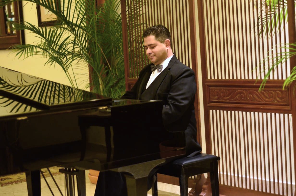 Yan Kayary de traje y moño, tocando un piano de cola en un lobby con biombo de madera y plantas
