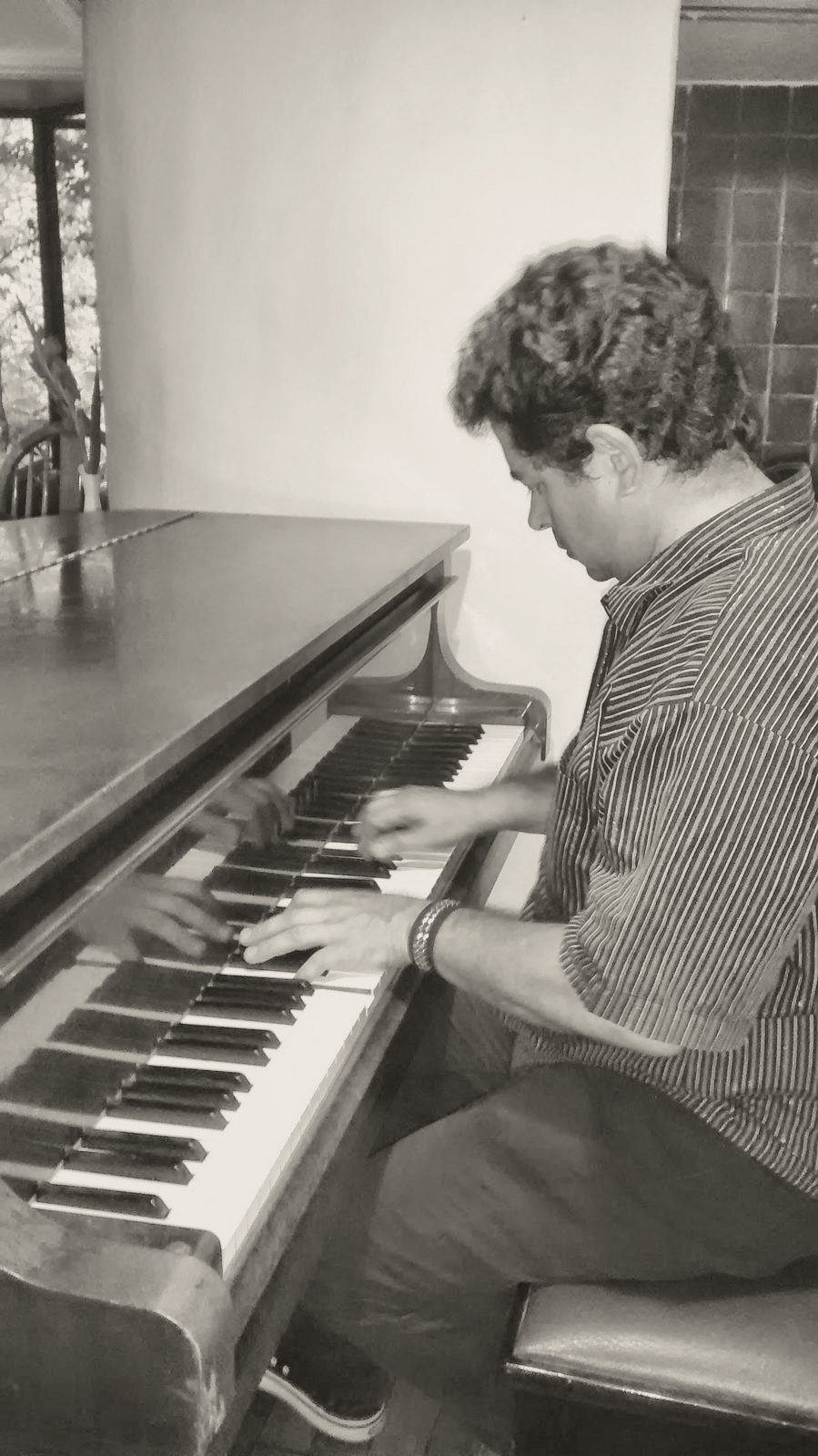 Yan Kayary con camisa azul, tocando un piano vertical de madera junto a una ventana